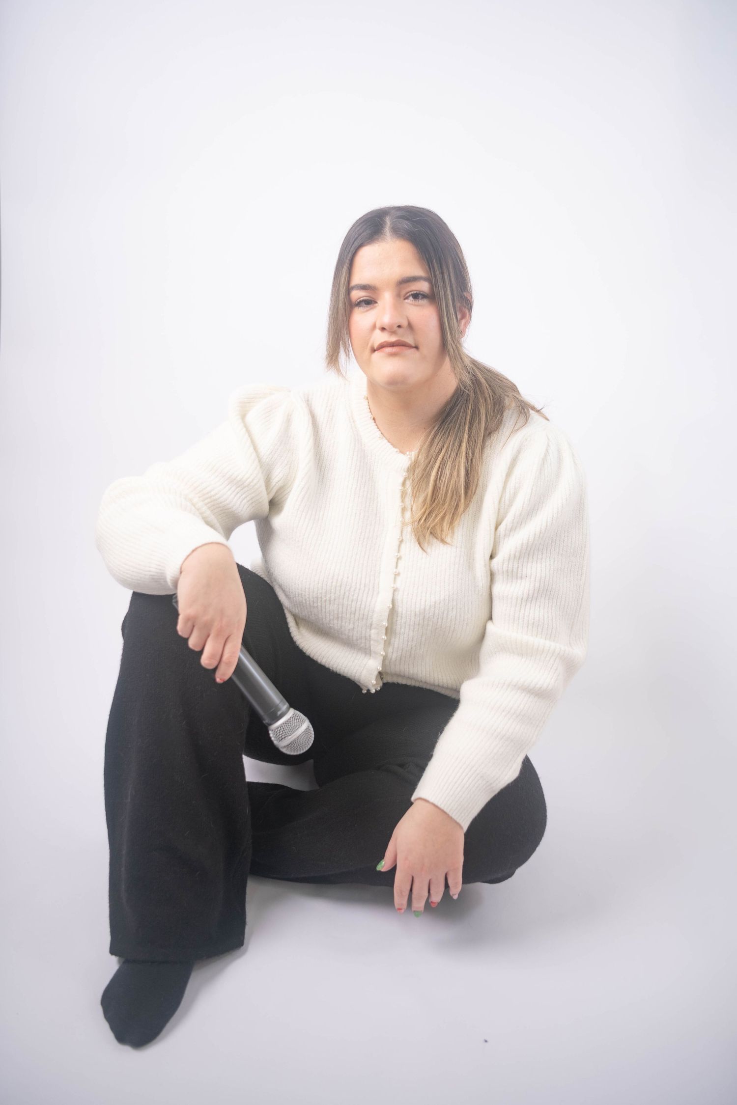 Young woman in white sweater sitting on floor holding microphone against white background.