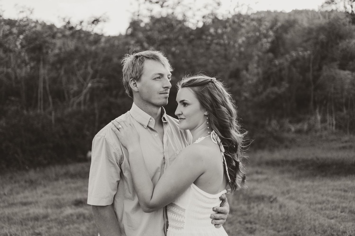 Black and white photo of a couple embracing in a field with trees in the background.