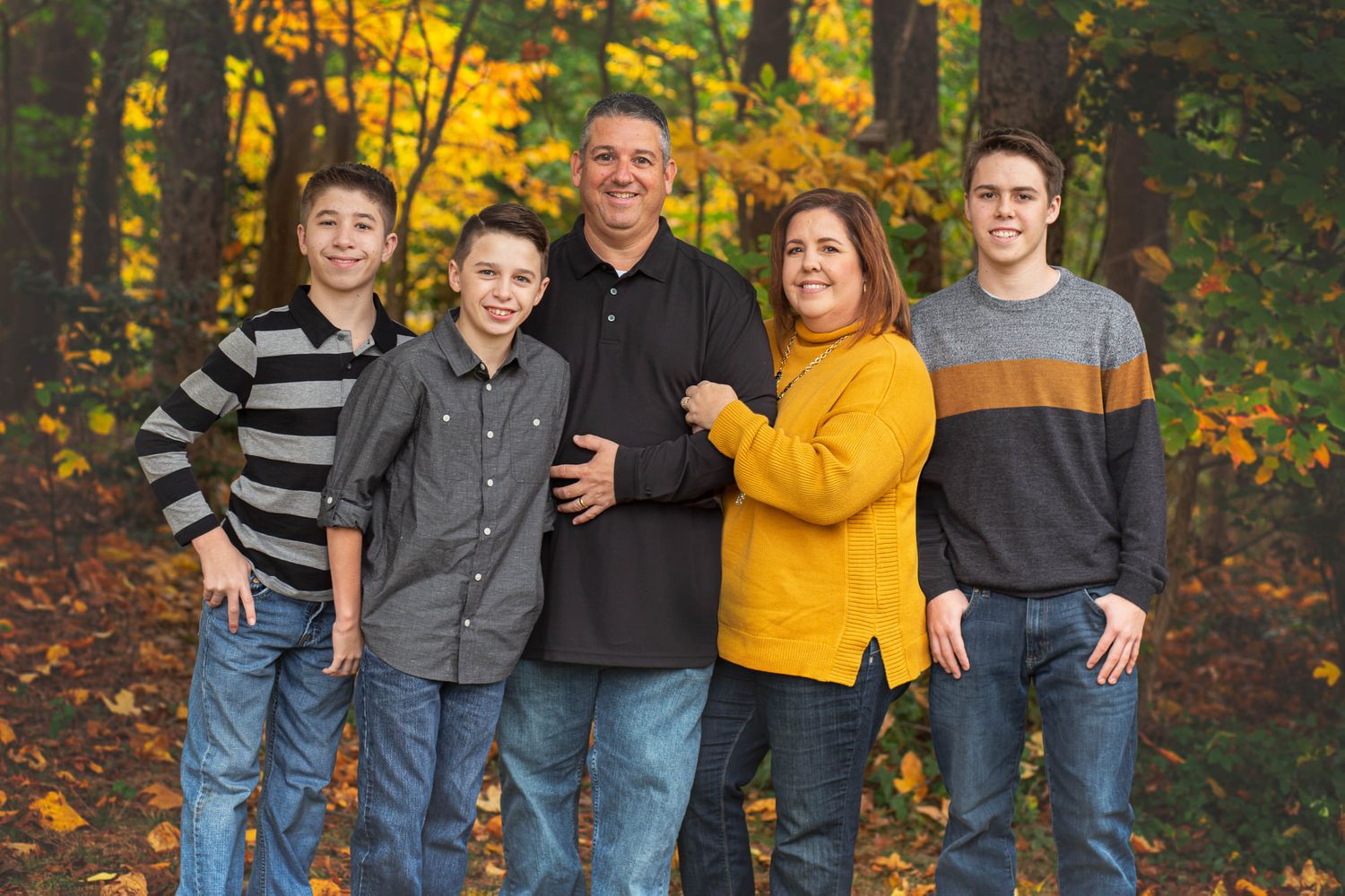 Family of five smiling together outdoors in autumn forest with colorful fall foliage backdrop.