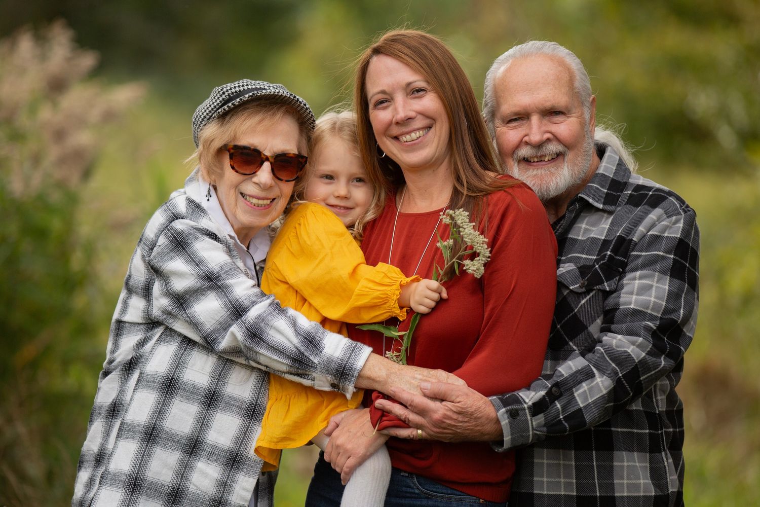 Family portrait of grandparents, mother, and young child embracing outdoors in a lush green setting.