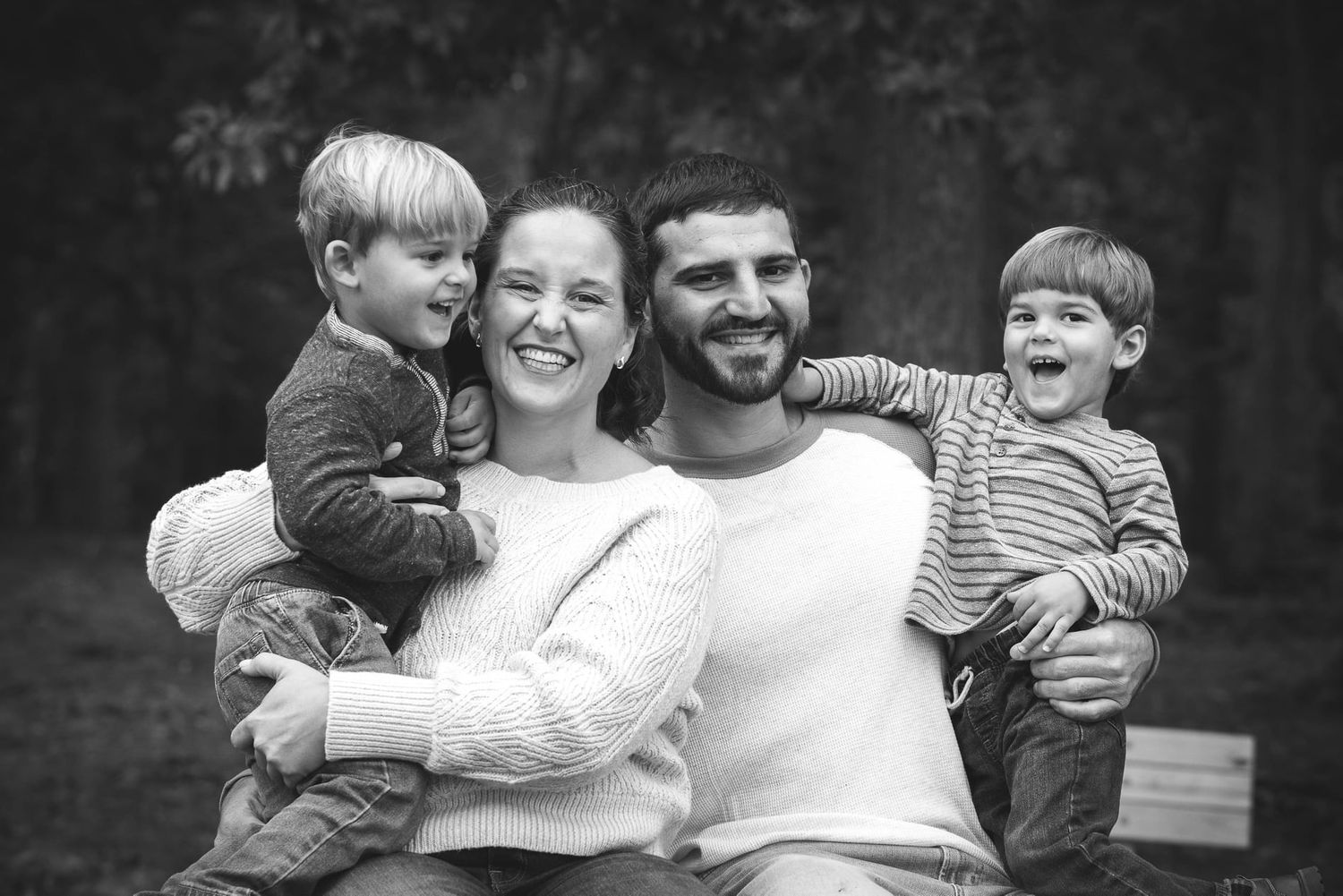Black and white family portrait of smiling parents holding two young boys outdoors in a wooded setting.