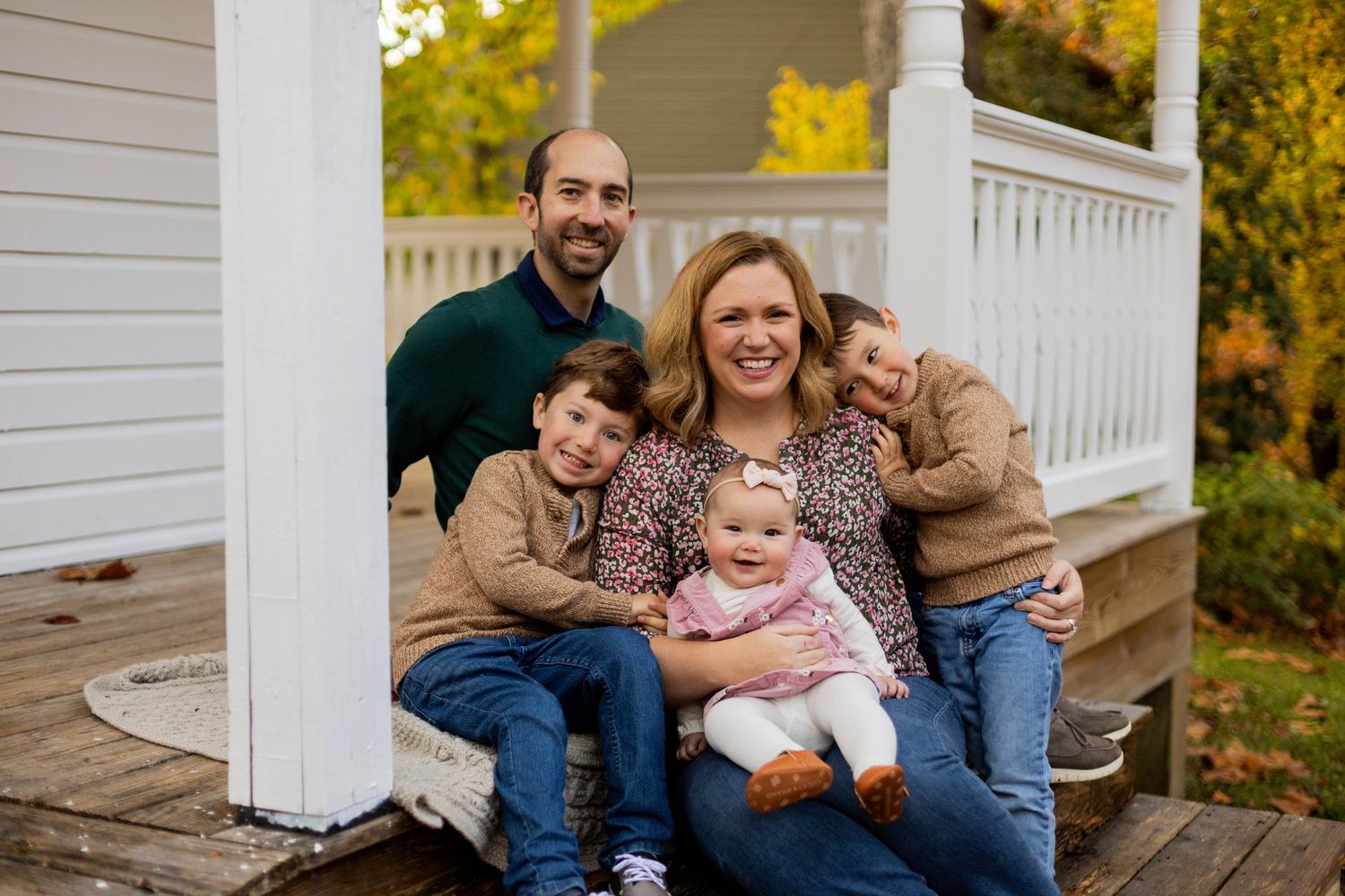 Happy family of five posing on porch steps in autumn, parents with three young children smiling outdoors.