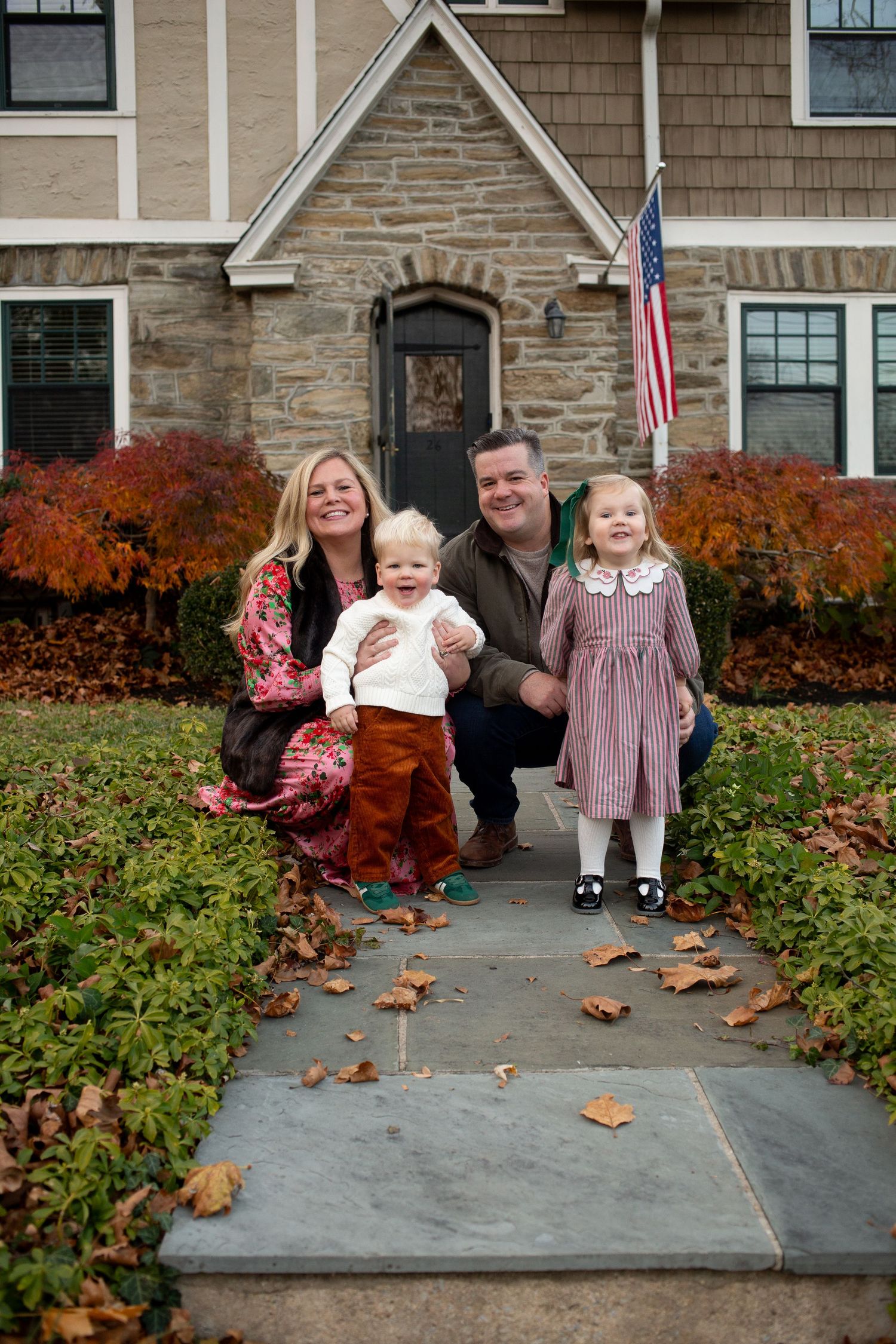 Family of four smiling in front of a stone house with fall foliage and an American flag on a autumn day.