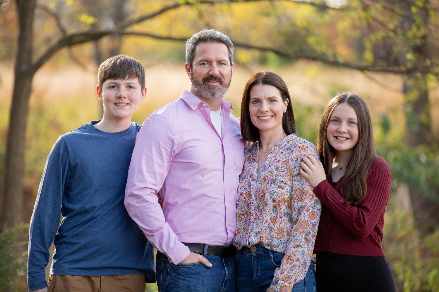 Family of four smiling outdoors in autumn park setting with colorful fall foliage in background.