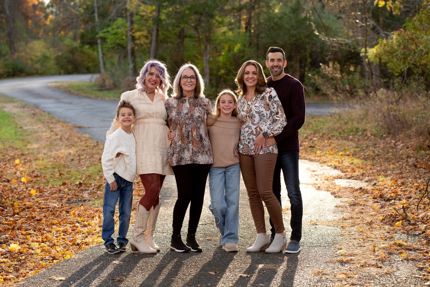 Multi-generational family of six smiling together on a tree-lined path surrounded by colorful autumn foliage.