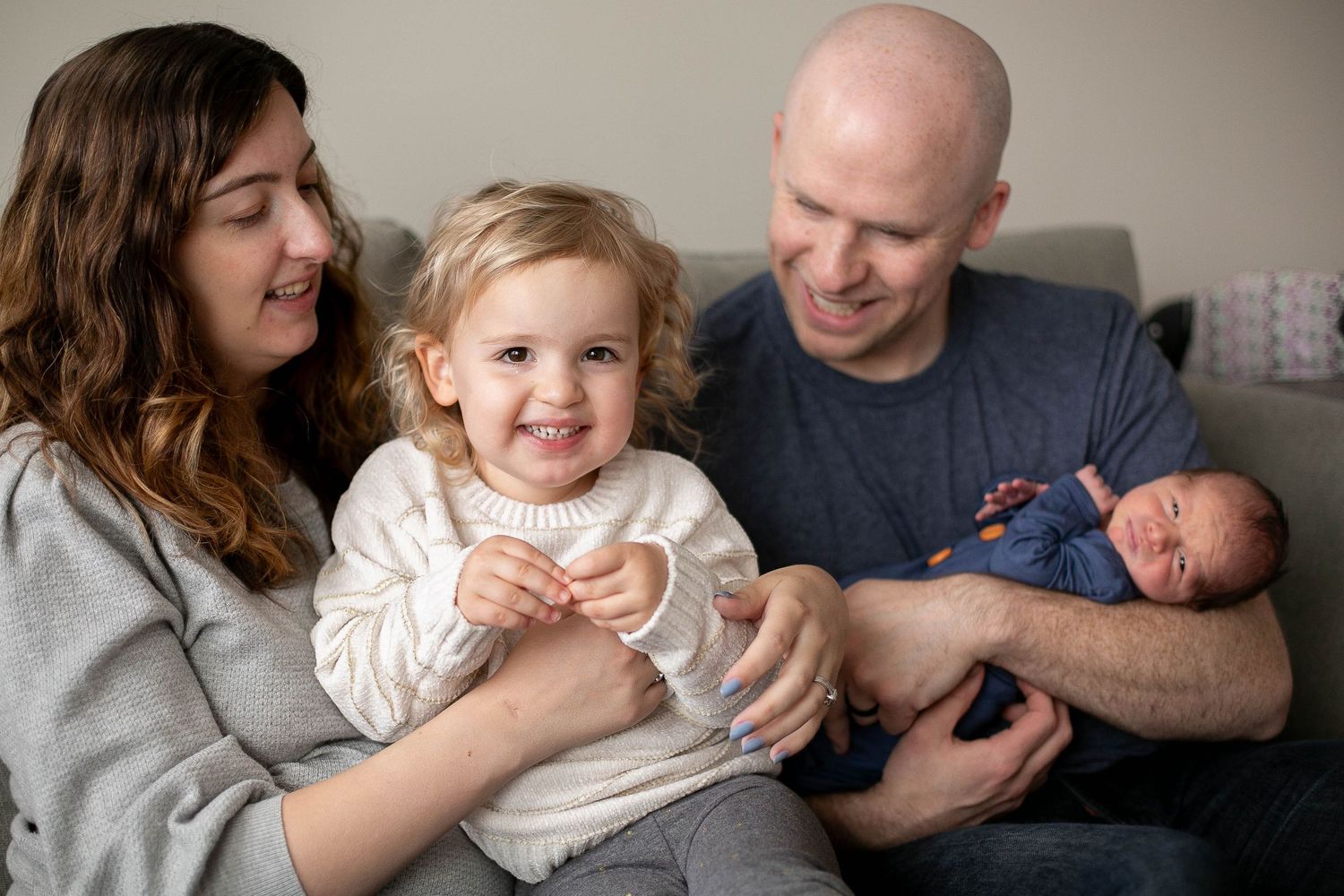 Happy family with toddler girl and newborn baby sitting together on a couch in a warm home setting.