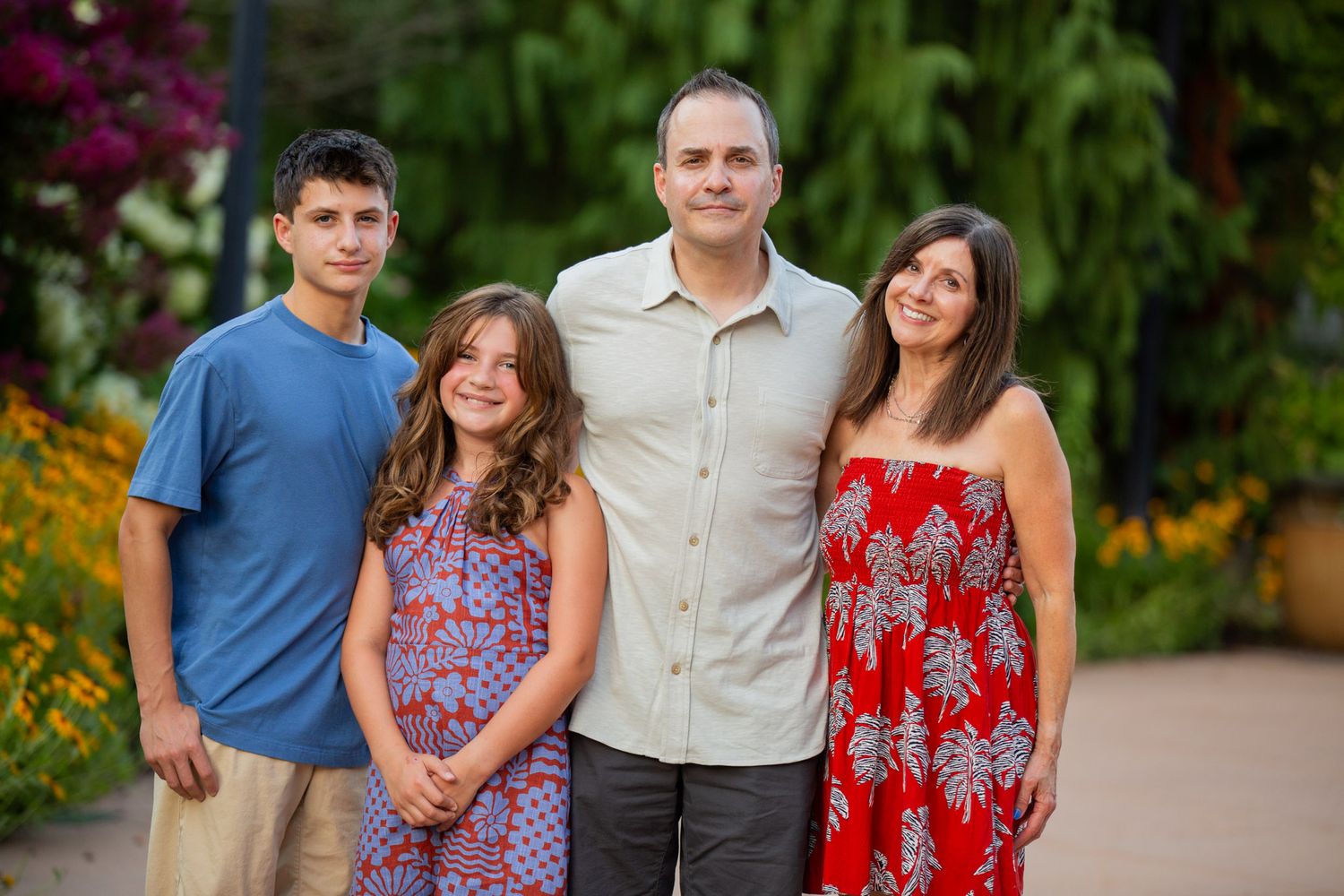 Family of four smiling outdoors in a lush garden setting, with colorful flowers in the background.