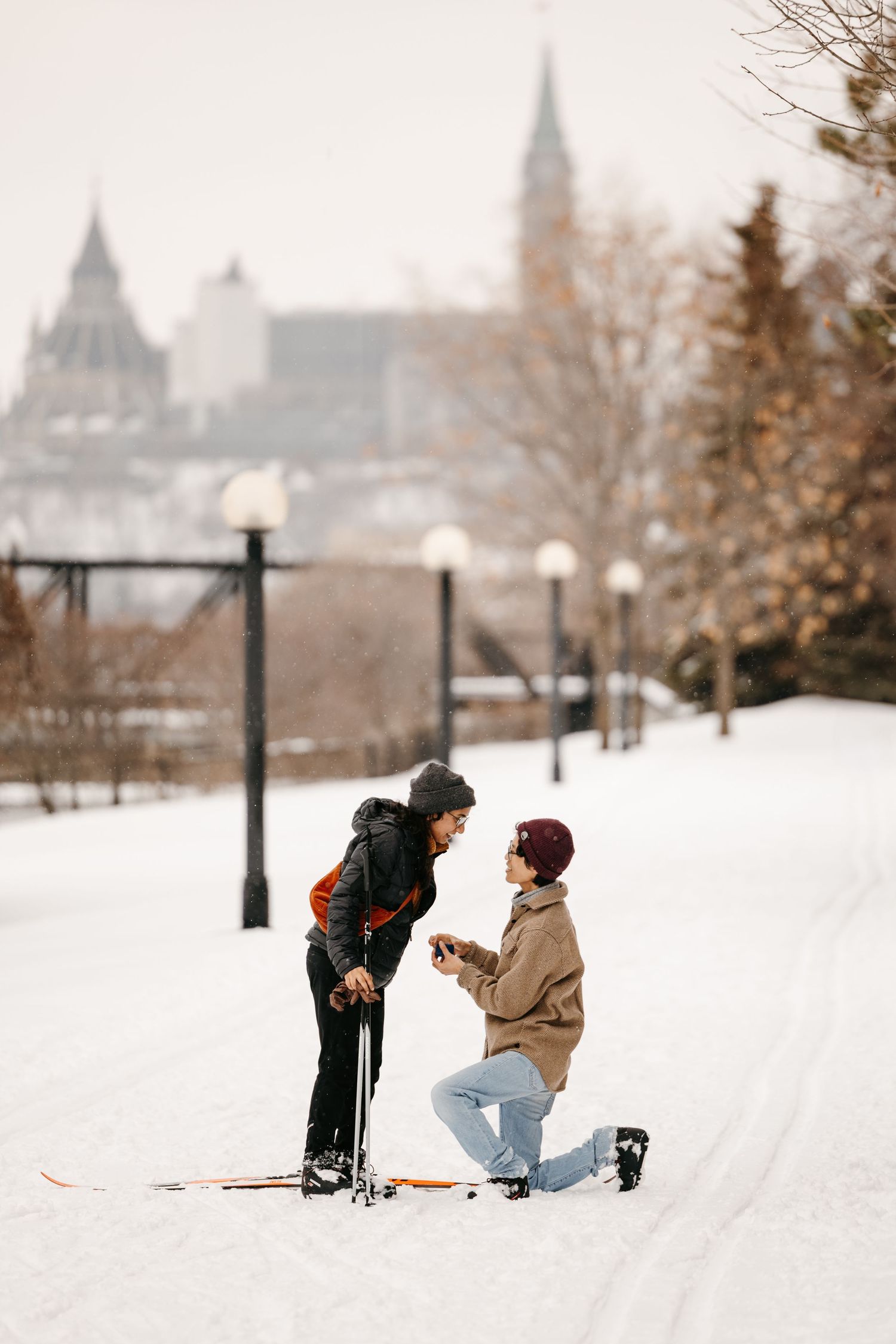 Romantic winter marriage proposal on snowy path with ski equipment, historic building backdrop in Ottawa, Canada.
