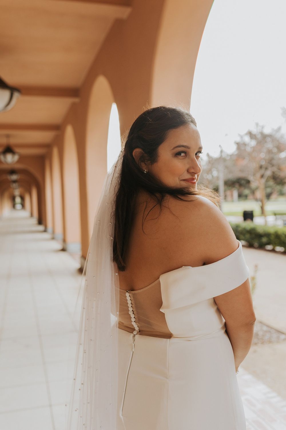 A bride in an elegant off-shoulder white wedding dress poses in a covered walkway with arched columns at Liberty Station.