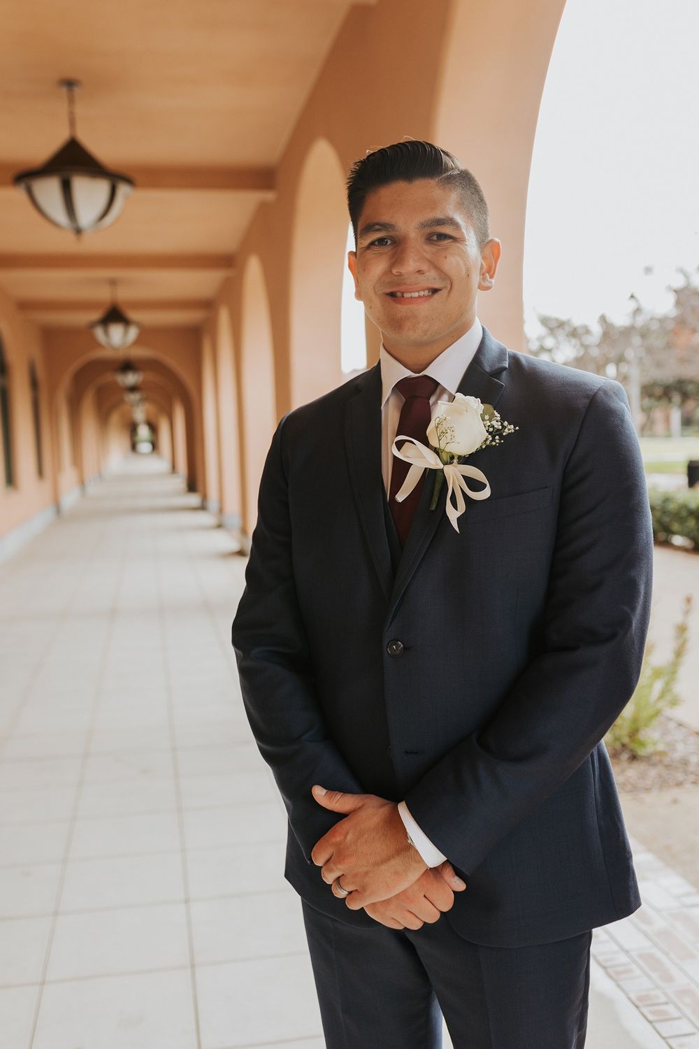 A groom in a black tuxedo smiles warmly while standing in an elegant covered walkway with arched columns at Liberty Station.