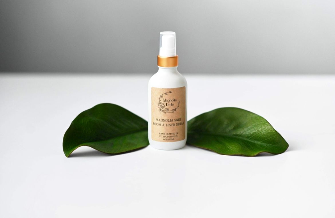 White spray bottle with gold cap surrounded by tropical green leaves on a clean white surface.