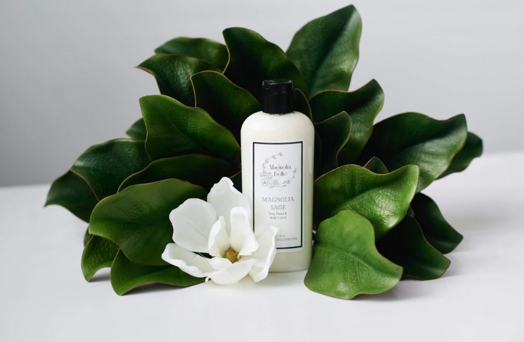 White lotion bottle surrounded by lush magnolia leaves and a white magnolia flower on a clean white surface.