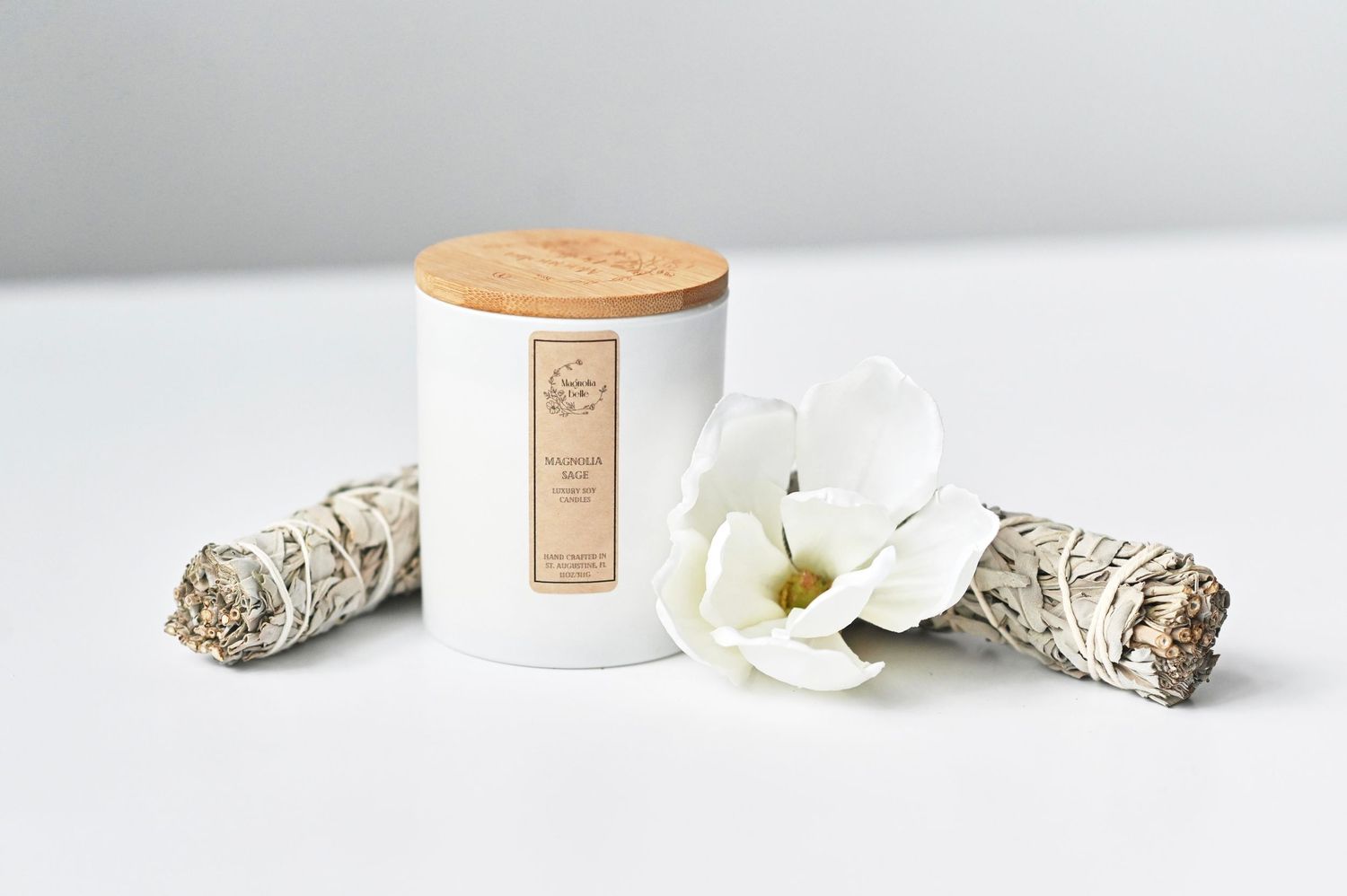 White candle with wooden lid beside sage bundles and white magnolia flower on clean white surface.