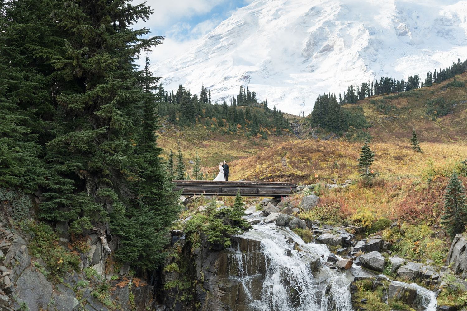 Two figures stand on a wooden bridge over cascading waterfalls with evergreen trees and mountain scenery in Mount Rainier.