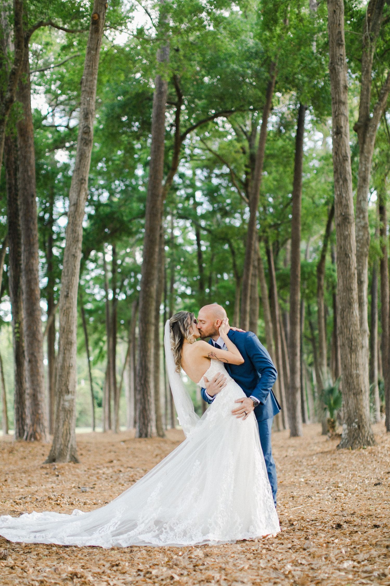 Bride and groom share a romantic and dramatic kiss among tall trees, bride in lace gown with veil, groom in navy suit in Bluffton, SC.