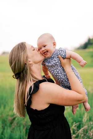 A woman in a black dress lifts and kisses a baby wearing a floral outfit in a sunny field.