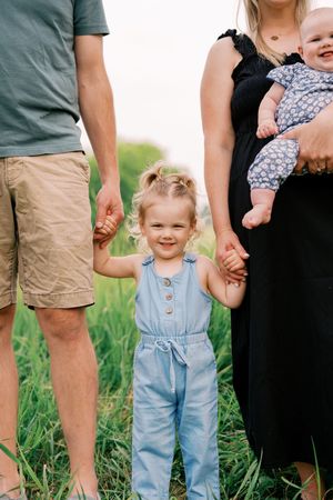 A family stands together in a grassy field, with a child in light blue overalls holding hands with parents.