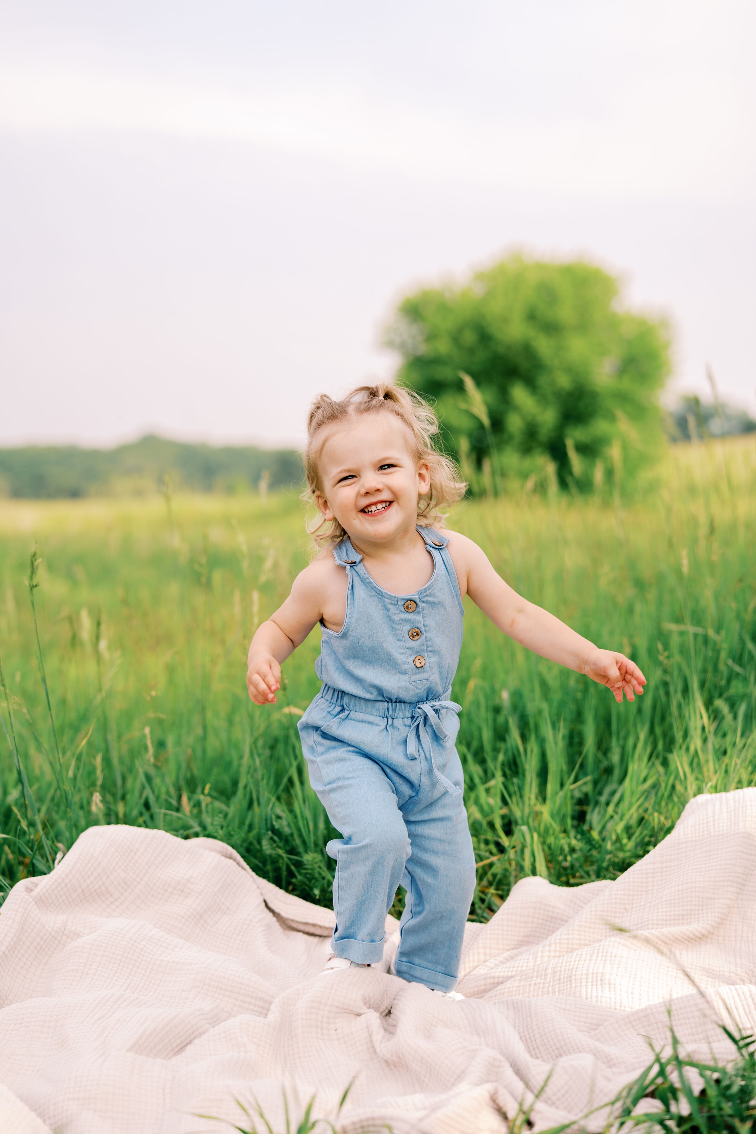 A joyful toddler in denim overalls plays on a blanket in a sunny meadow with green trees in the background.