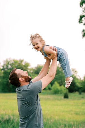 Parent and child share joyful moments playing and lifting in a sunny field with green grass.