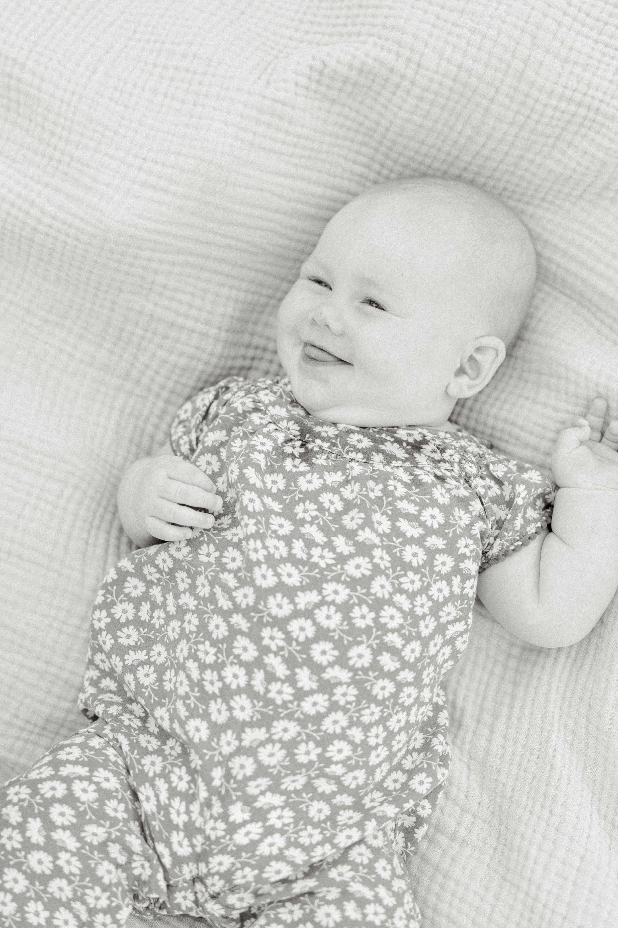 A baby in a floral print onesie lies peacefully on white bedding in a black and white photograph.
