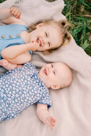 Two smiling children laying on a cream colored blanket in grass on a sunny day.