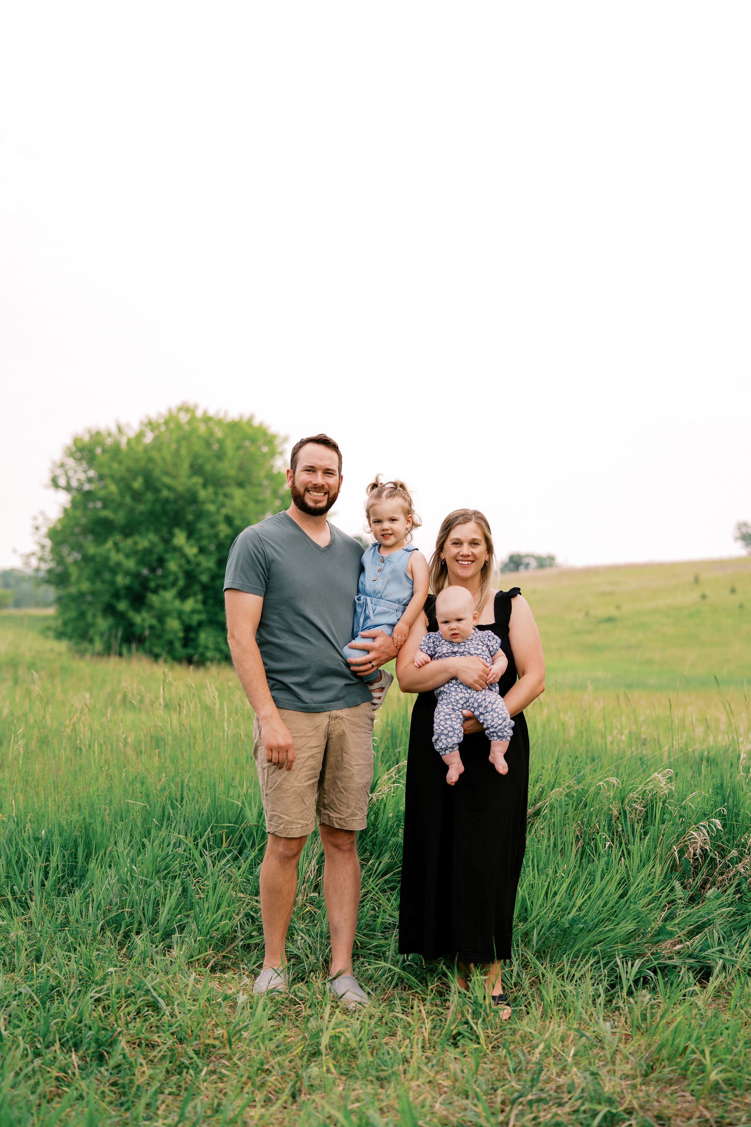A family stands together in a green meadow on a sunny day, with tall grass and trees in the background.