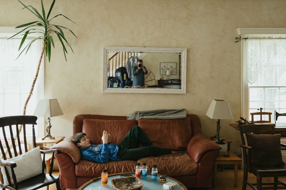 A panoramic view of a living room with brown leather couch, houseplants, and natural lighting from windows.
