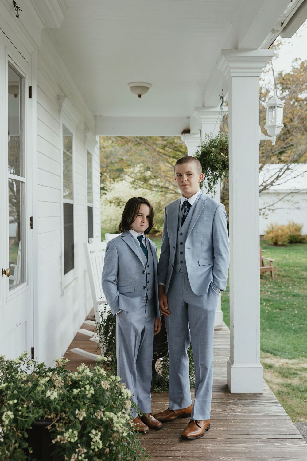 Wedding party members in light gray suits pose on a white columned porch of a rustic farmhouse.