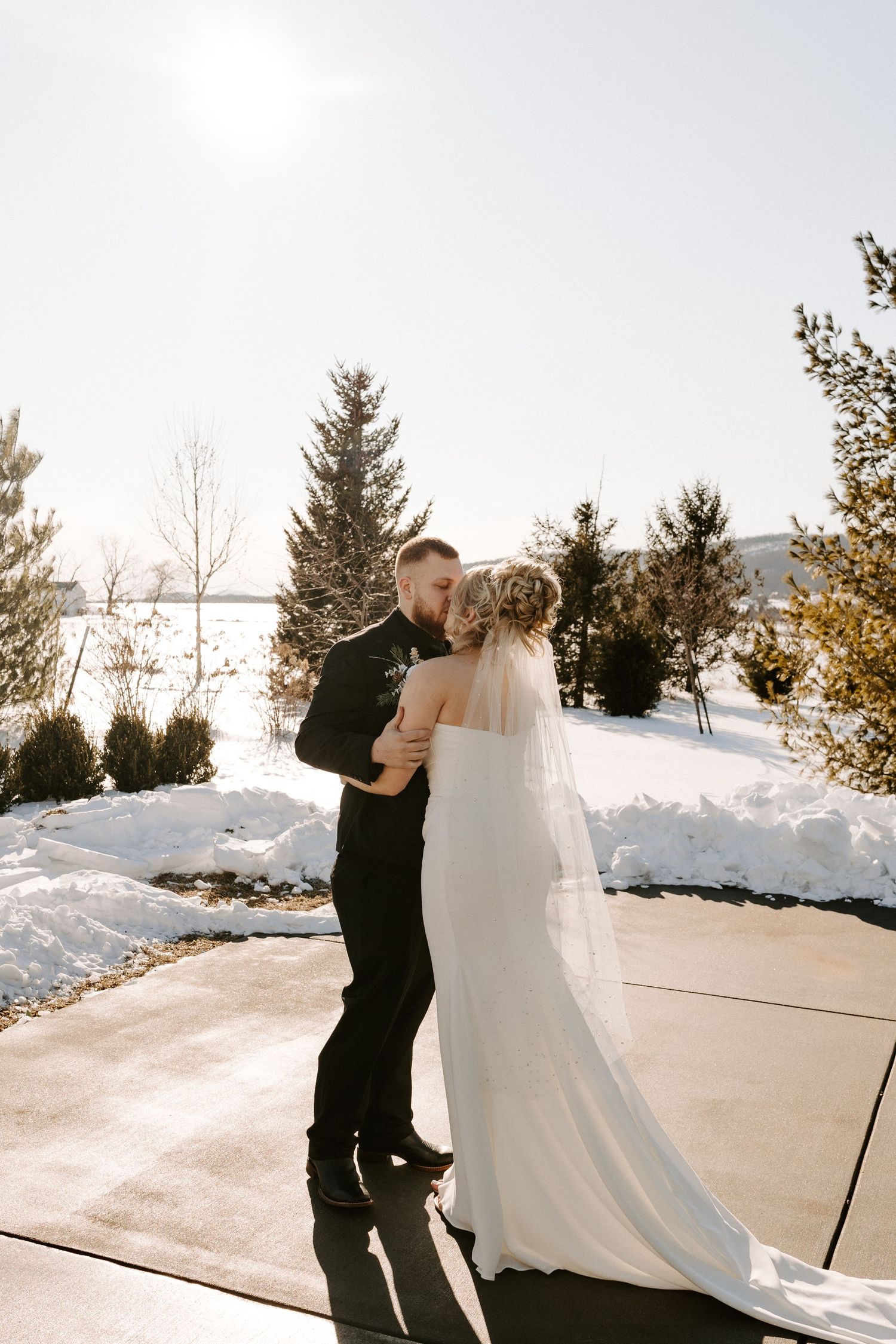 Bride and groom share first kiss outdoors in snowy winter wedding setting with evergreen trees behind them.