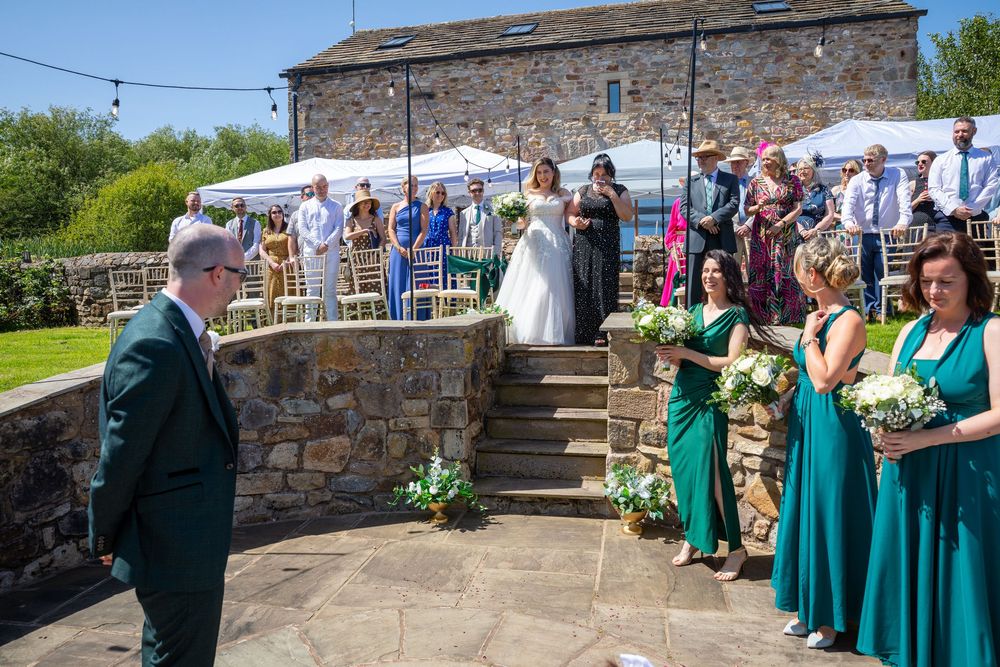 Bride descends stone steps toward groom as bridesmaids in teal dresses look on outdoors.
