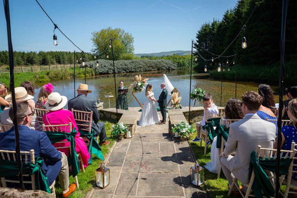 Outdoor wedding ceremony by a lakeside with guests seated on gold chairs in bright sunshine.