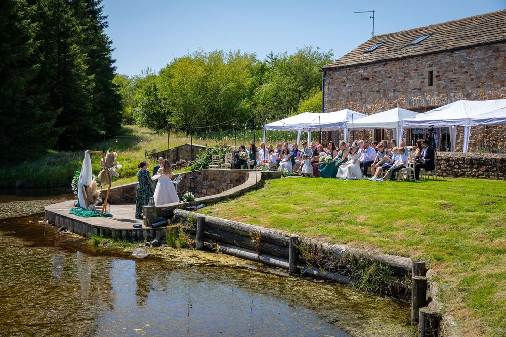 Outdoor wedding ceremony by a pond at a rustic stone barn venue on a sunny day.