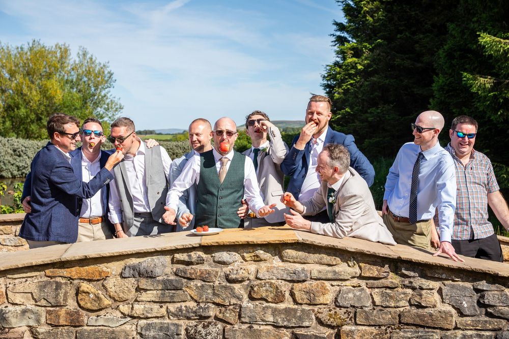 Group of groomsmen laughing and posing together at a stone wall on a sunny wedding day.