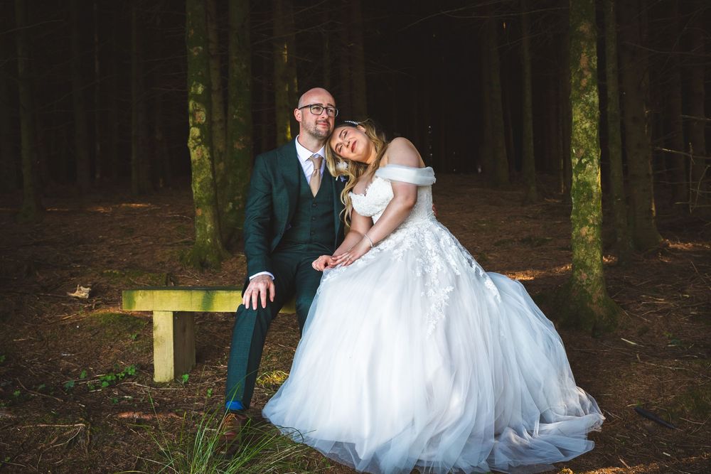 Bride and groom sharing a romantic moment on a bench surrounded by tall trees in a forest.