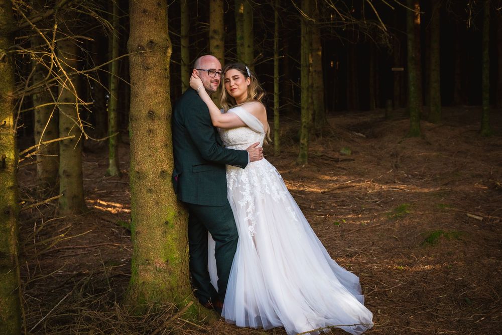 Bride and groom embracing against a tree trunk in a moody forest wedding portrait.