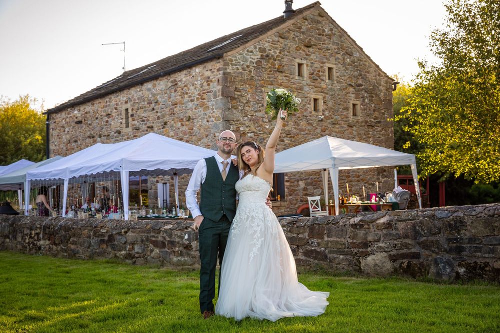 Bride raising bouquet triumphantly with groom outside rustic stone barn wedding venue at dusk.