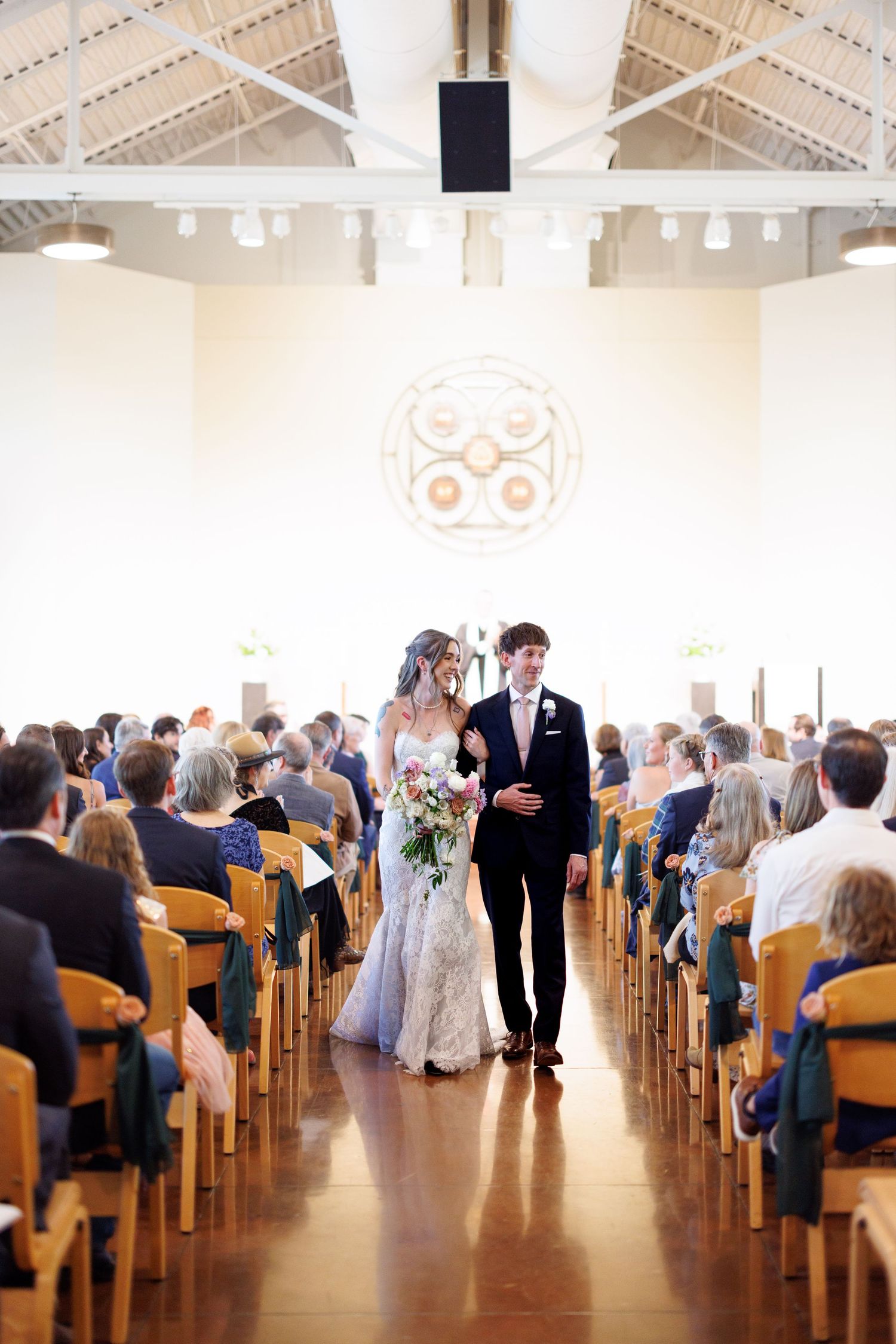 A panoramic view of a wedding ceremony in a bright church hall with guests seated in wooden pews.