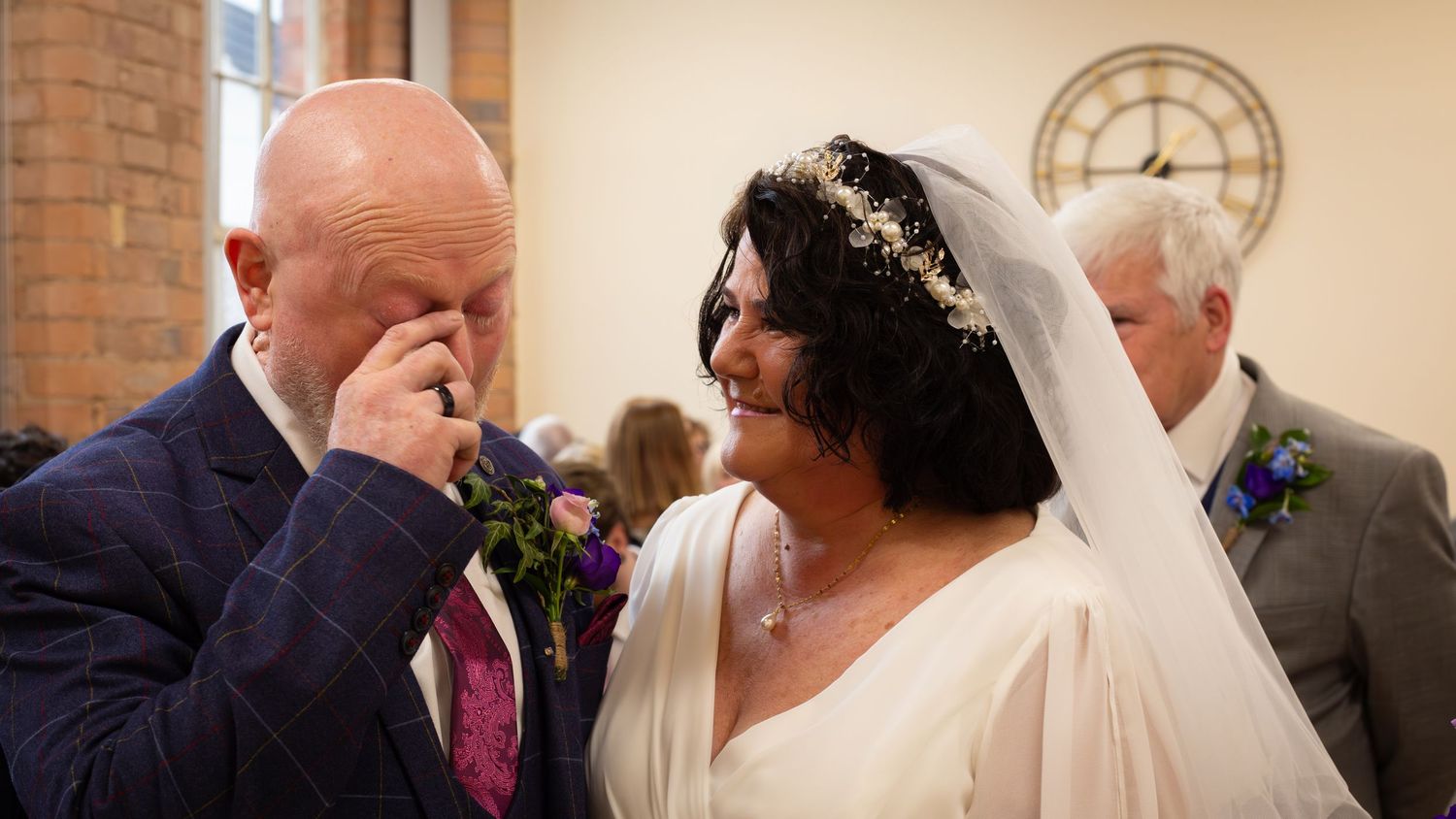 Emotional groom wipes tears as bride smiles in white gown and floral crown during wedding ceremony in church.