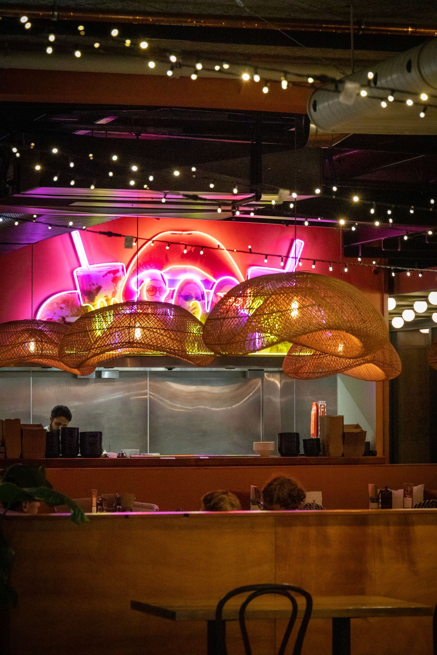 Vibrant restaurant interior with woven lampshades, neon signage, and fairy lights creating a lively dining scene.