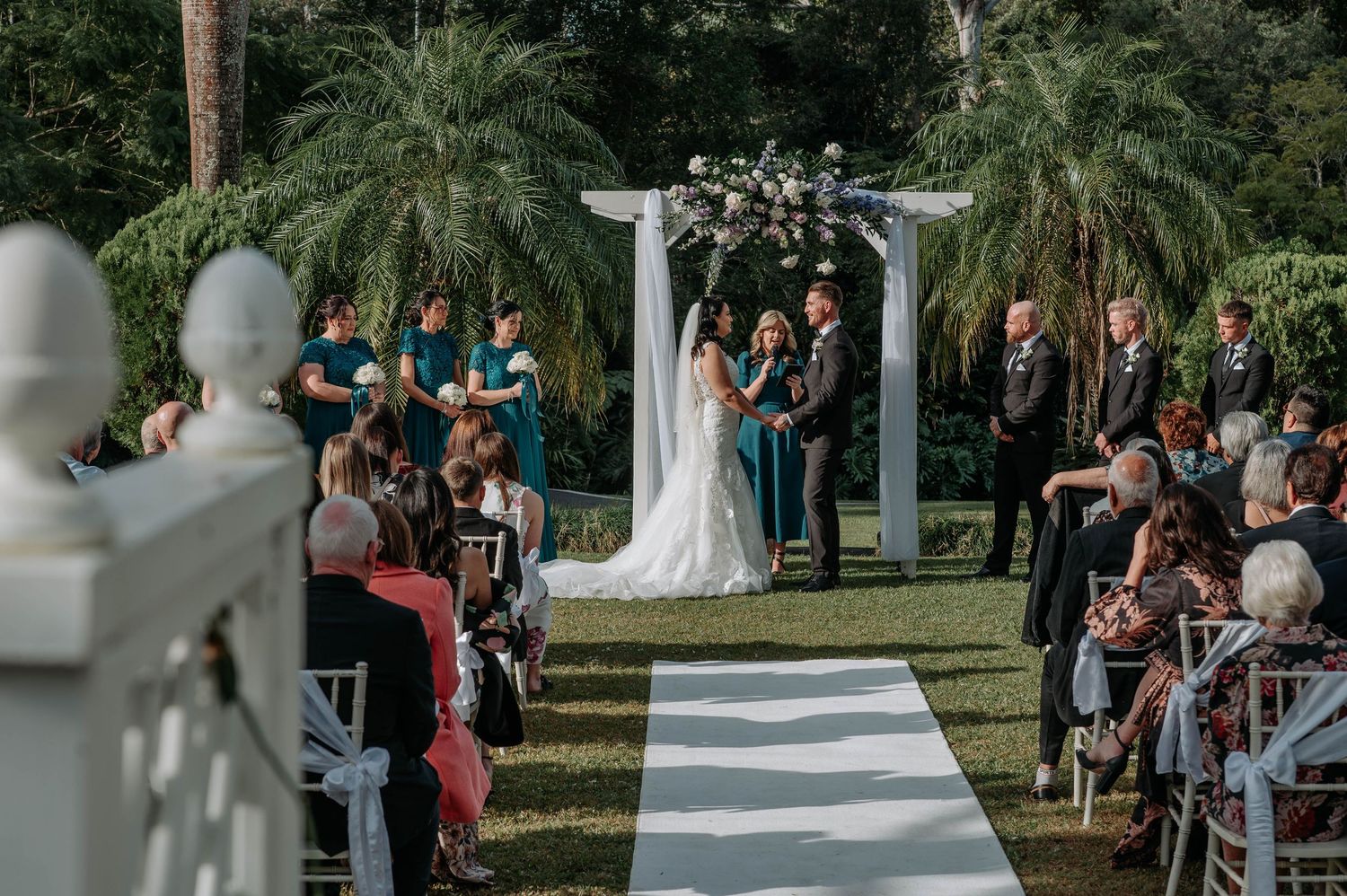 An outdoor wedding ceremony under a white pergola with palm trees and guests seated on both sides of a striped aisle runner.