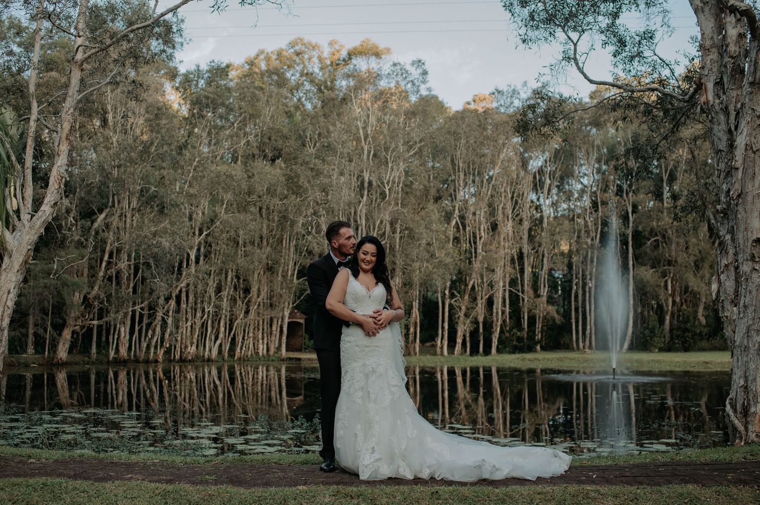 A couple in wedding attire stands in front of a scenic wooded area at dusk creating a romantic silhouette.