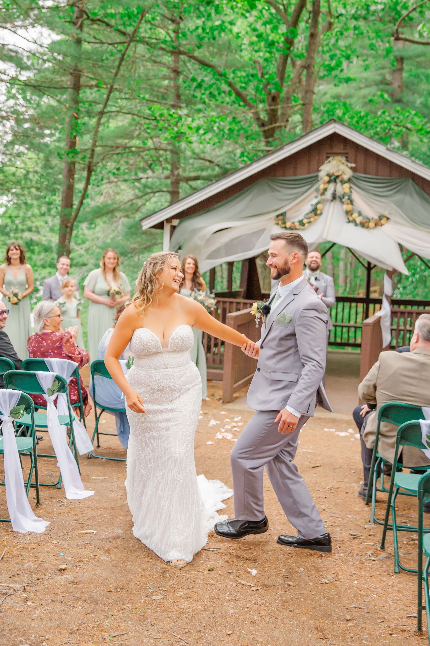 A couple dances joyfully at their outdoor wedding ceremony under a rustic wooden pavilion decorated with floral garlands.