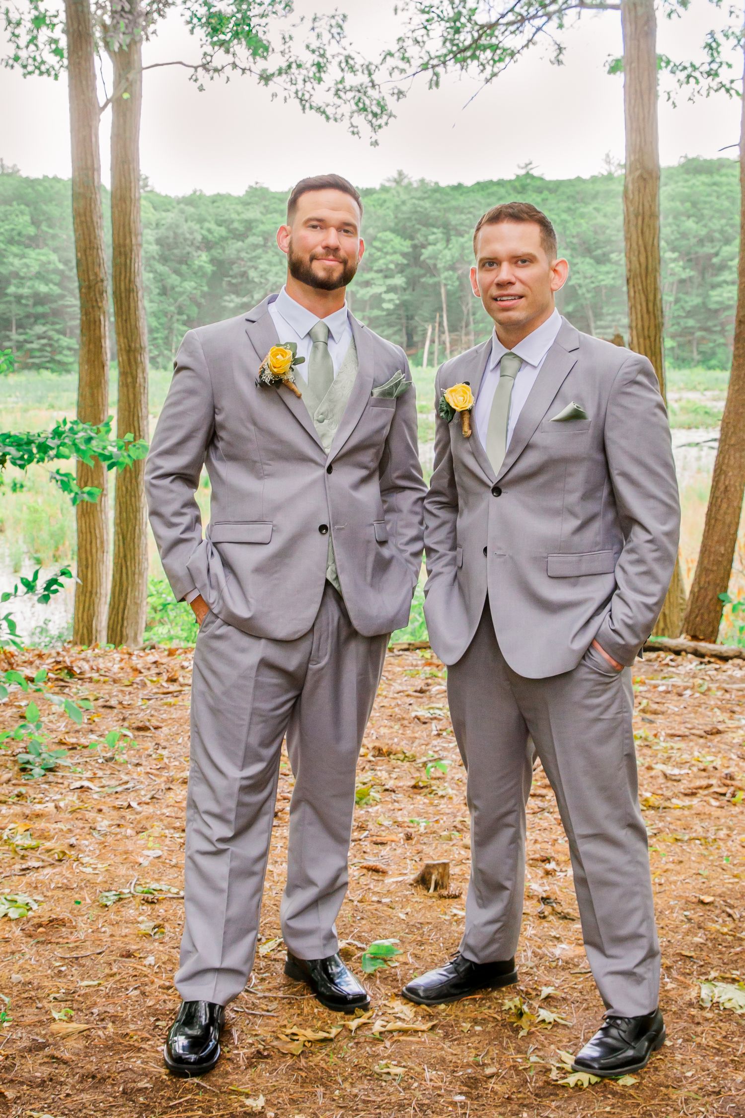 Wedding party members in matching light gray suits with yellow boutonnieres pose together in a wooded outdoor setting.