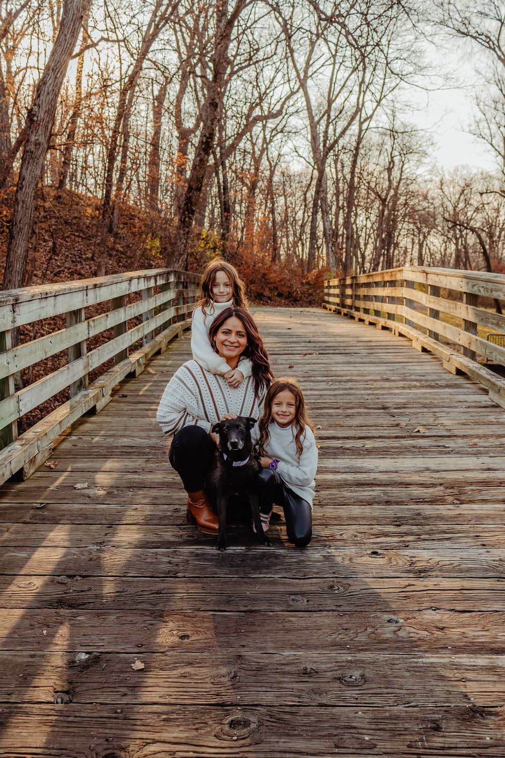 A small group poses with their black dog on a wooden boardwalk surrounded by autumn trees at sunset.