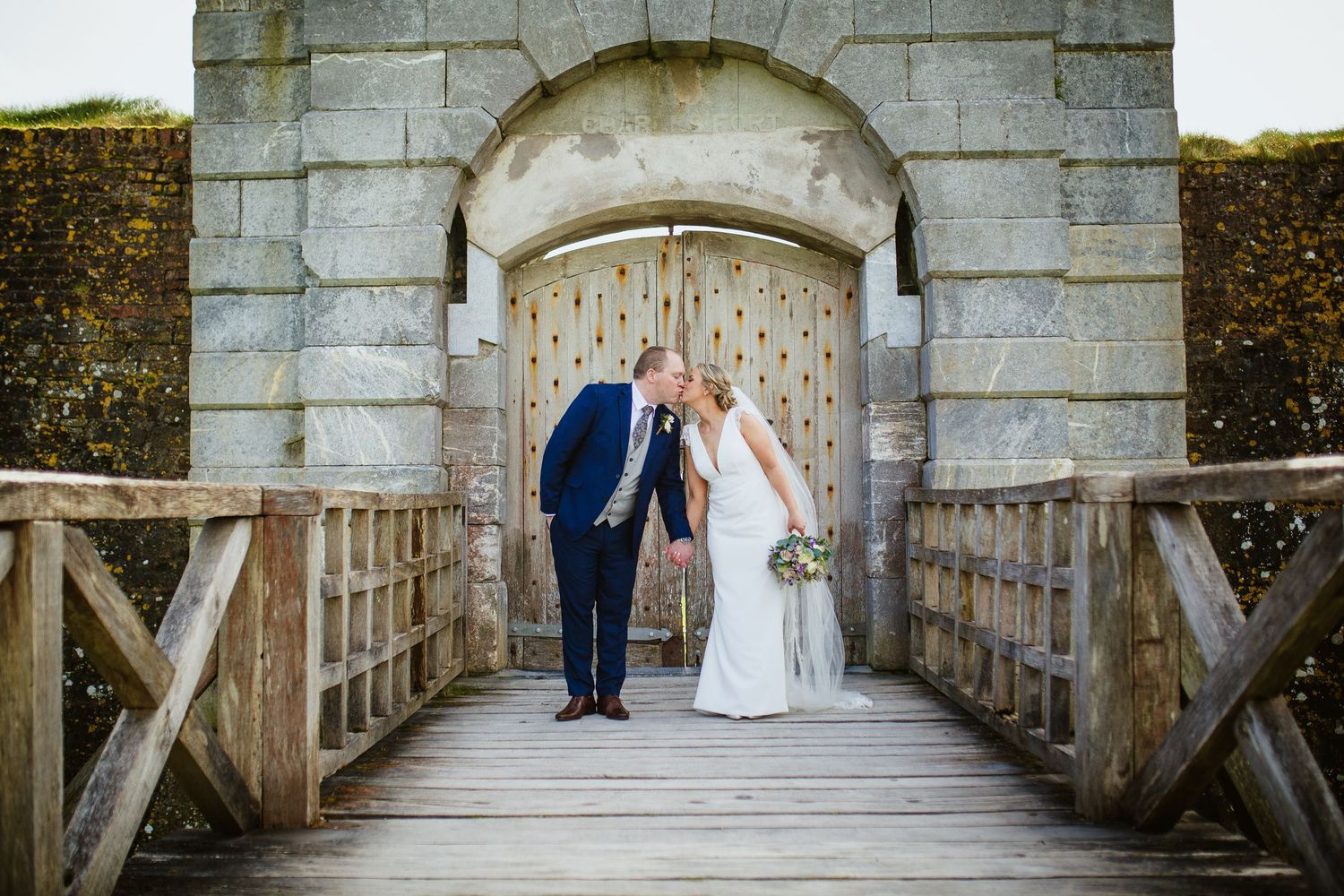A couple shares an intimate moment on a wooden bridge beneath a stone archway at a historic venue.