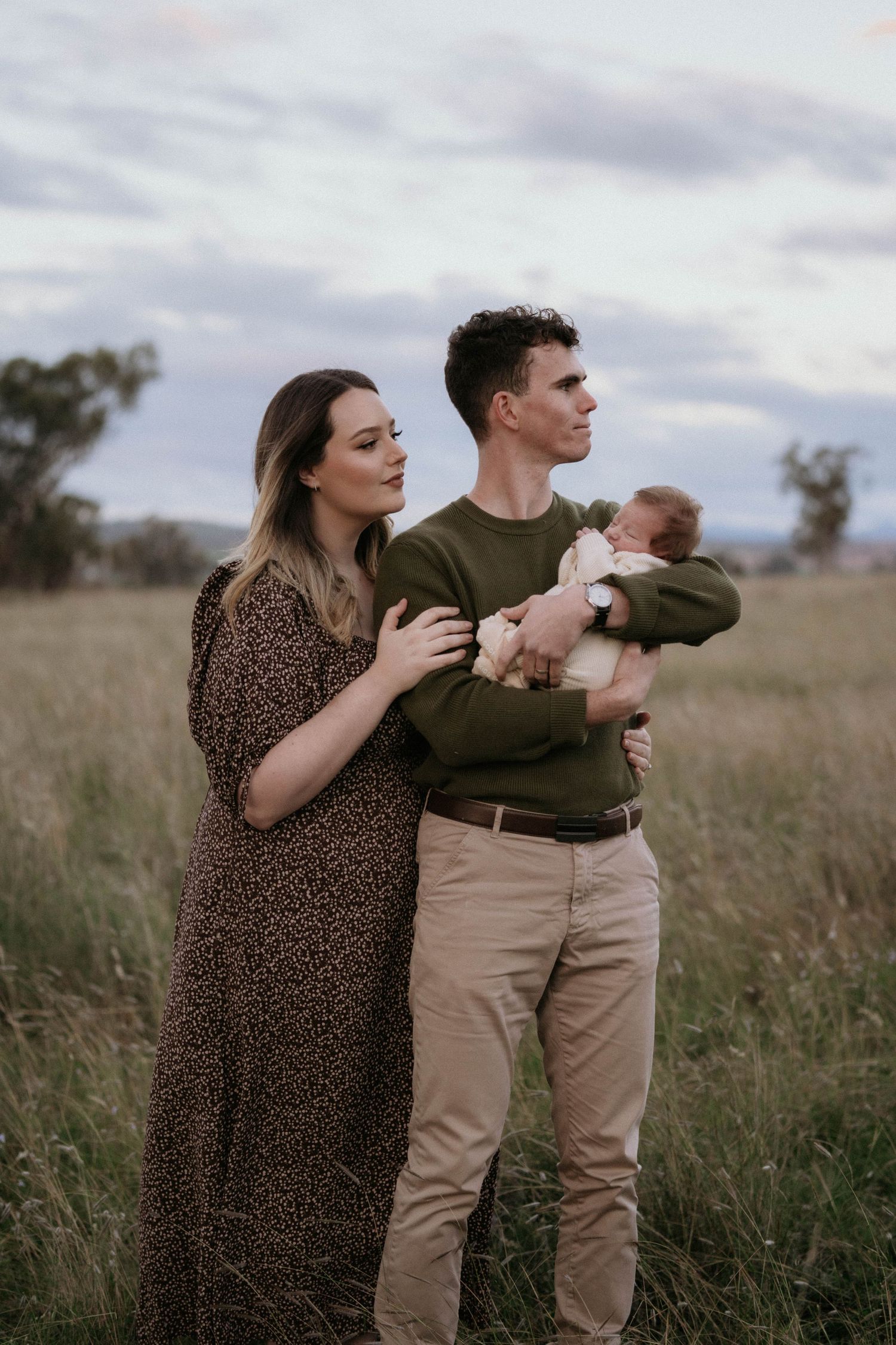 Couple stands together in a grassy field holding their newborn baby during golden hour with moody cloudy sky.