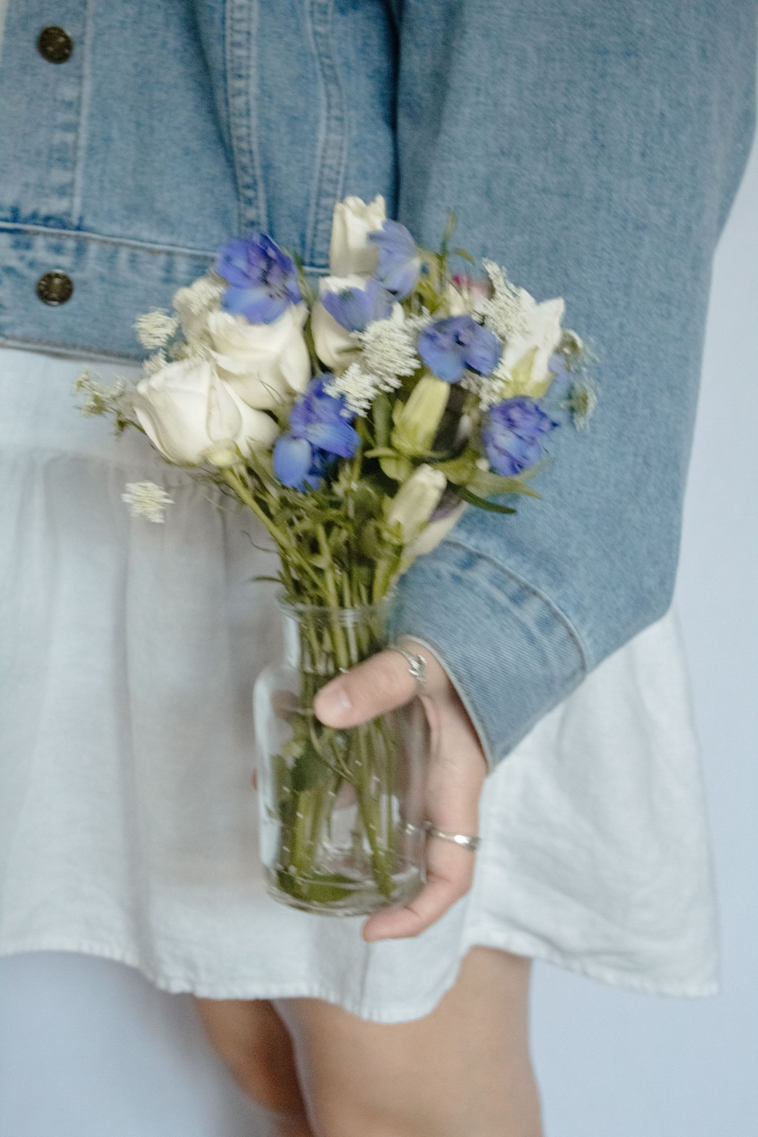 Hand holding a mason jar with delicate white roses and purple wildflowers against denim and white fabric background.