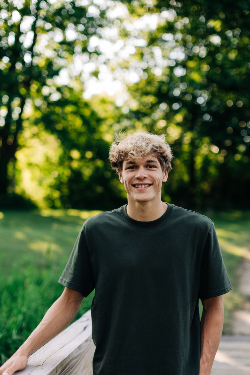A person in a dark t-shirt smiles while standing on a path with green trees and bokeh lighting in the background.