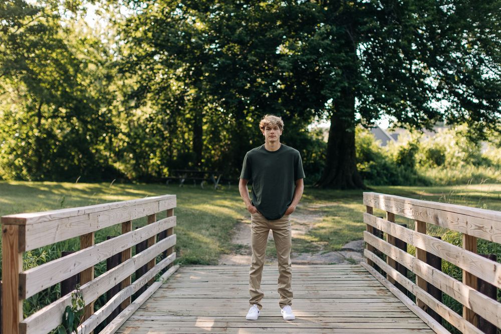 A figure stands centered on a wooden bridge walkway surrounded by lush green trees in summertime lighting.