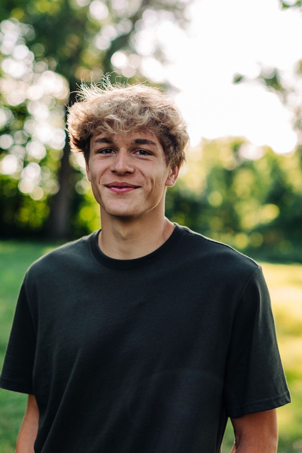 A smiling individual in a black t-shirt stands outdoors with blurred trees and natural lighting in the background.